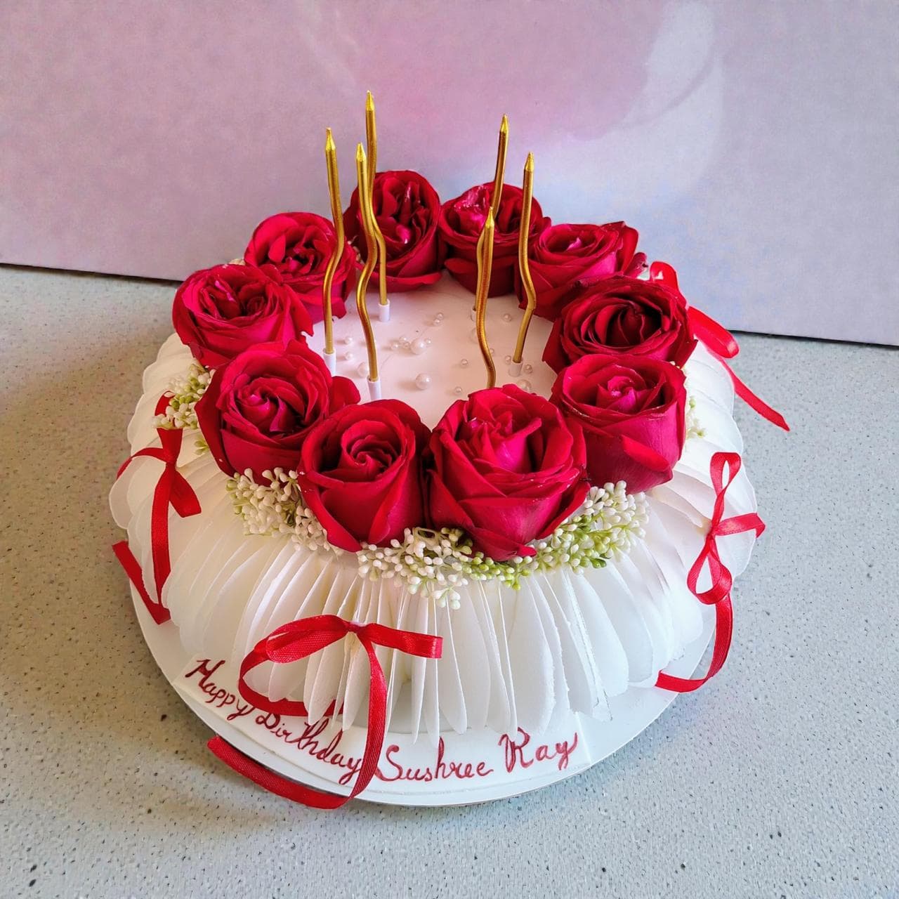 White celebration cake topped with red roses, gold candles, and ribbon details